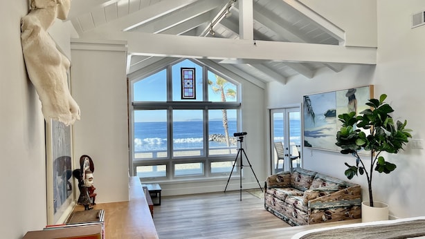 Un'ampia stanza con una grande finestra vista spiaggia, soffitto alto con travi a vista, una fotocamera su un treppiede e un’accogliente zona salotto con un divano a motivi.