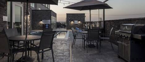 Un patio al aire libre con suelo de piedra, mesas y sillas de metal, sombrillas y una parrilla de barbacoa.