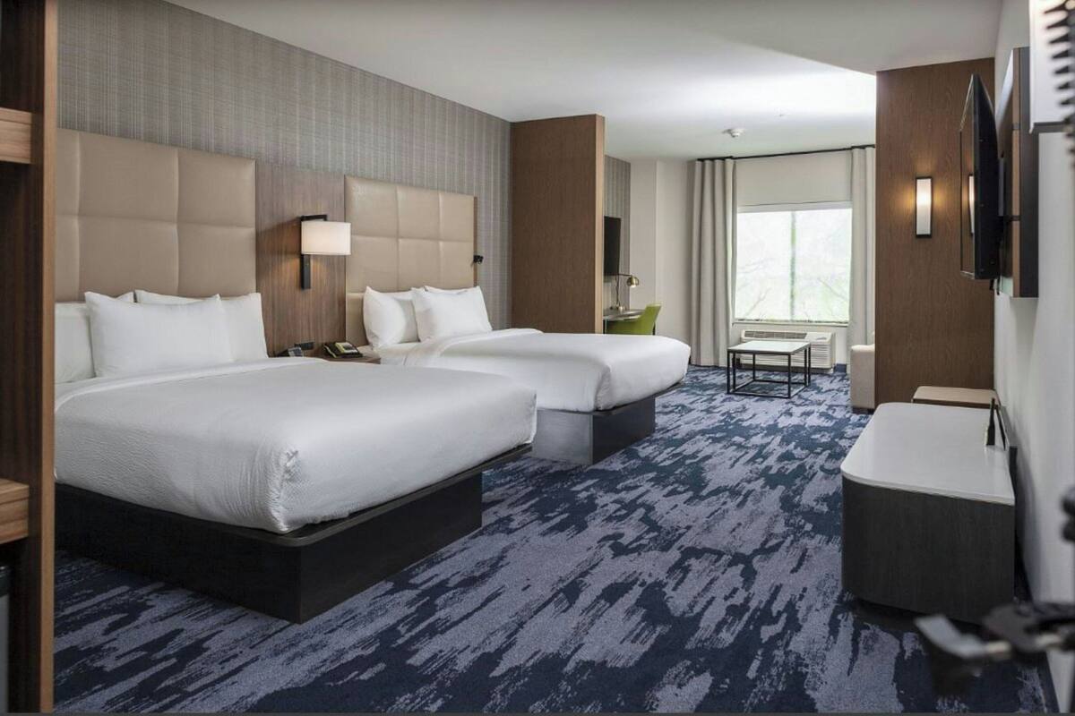 A hotel room with two beds, a desk, a chair, a TV, and a window.