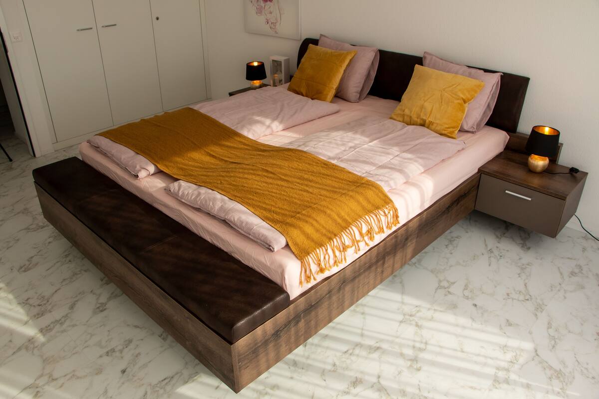 Una cama bien hecha con un cabecero marrón, una mesita de noche con lámpara y un armario al fondo.