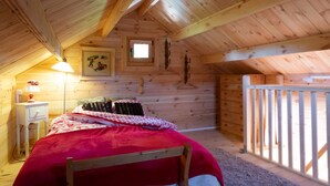 A cozy attic bedroom with a wooden ceiling, a small window, a bed with a red blanket, a bedside table with a lamp, and a view of the outside balcony.