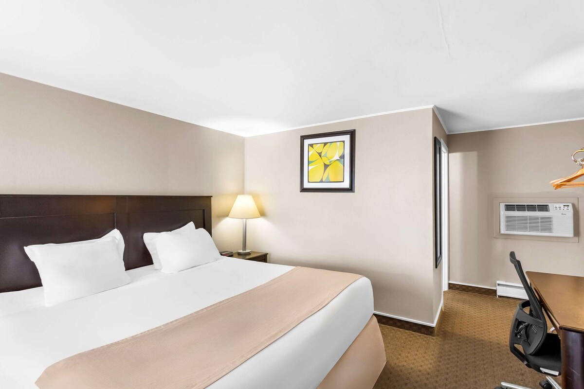 A hotel room with a bed, a nightstand with a lamp, a framed picture on the wall, and a desk with a chair.
