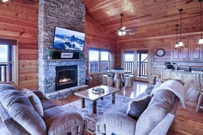 A cozy log cabin interior with a stone fireplace, comfortable seating, a television, and a kitchen area. - Panoramic View - Suite 4: Poker Table - Spectacular Mountain Backdrop (sevierville)