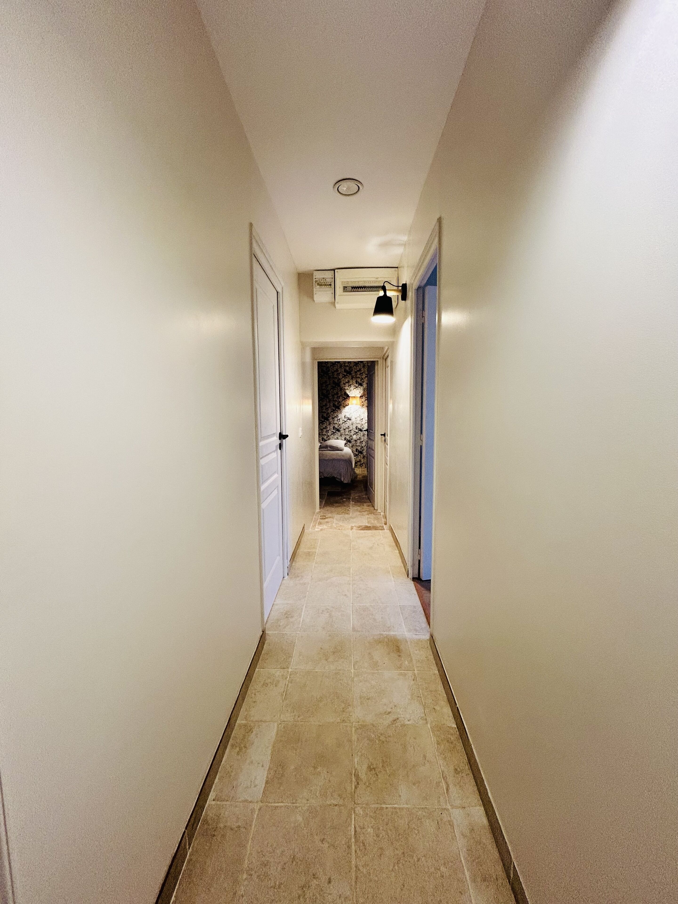 A narrow, well-lit hallway with tiled flooring, leading to a bedroom with a floral-patterned wall and a bed.