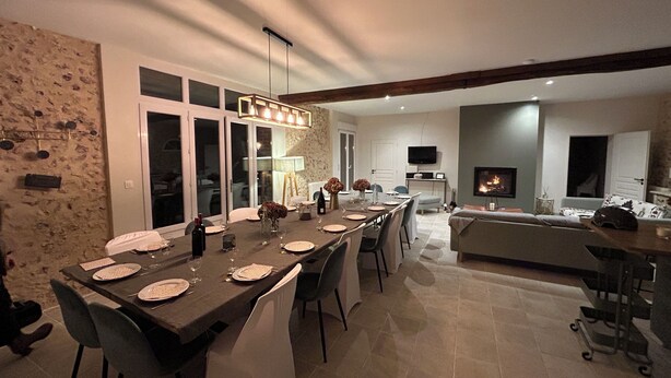 A modern kitchen with a dining area, featuring a long table set for six, a fireplace, and a television mounted on the wall.
