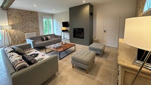 A modern living room with a grey sofa, a wooden coffee table, and a grey ottoman. There is a fireplace, a TV mounted on the wall, and a lamp on a side table.