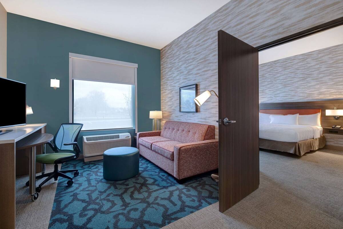 Quarto de hotel com cama, sofá, escrivaninha com computador, cadeira, luminária e janela.