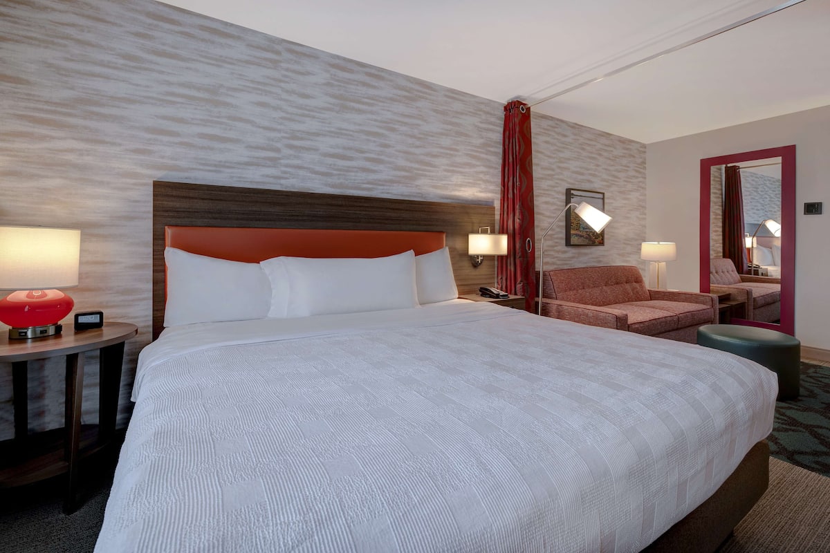 Hotelkamer met een groot bed, twee nachtkastjes met lampen, een rood gordijn, een spiegel en een zithoek met een fauteuil en poef.