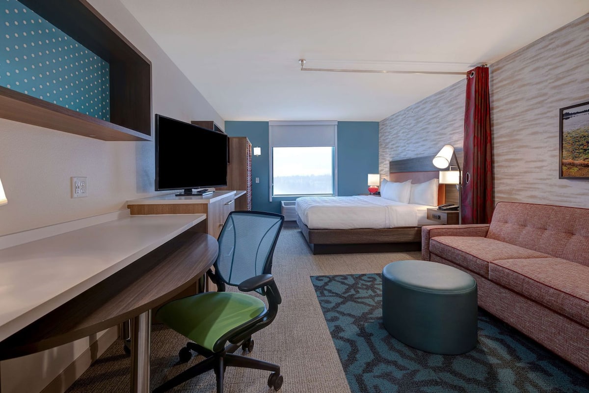 Quarto de hotel com cama, escrivaninha com televisão, cadeira de escritório verde, sofá vermelho e tapete estampado.
