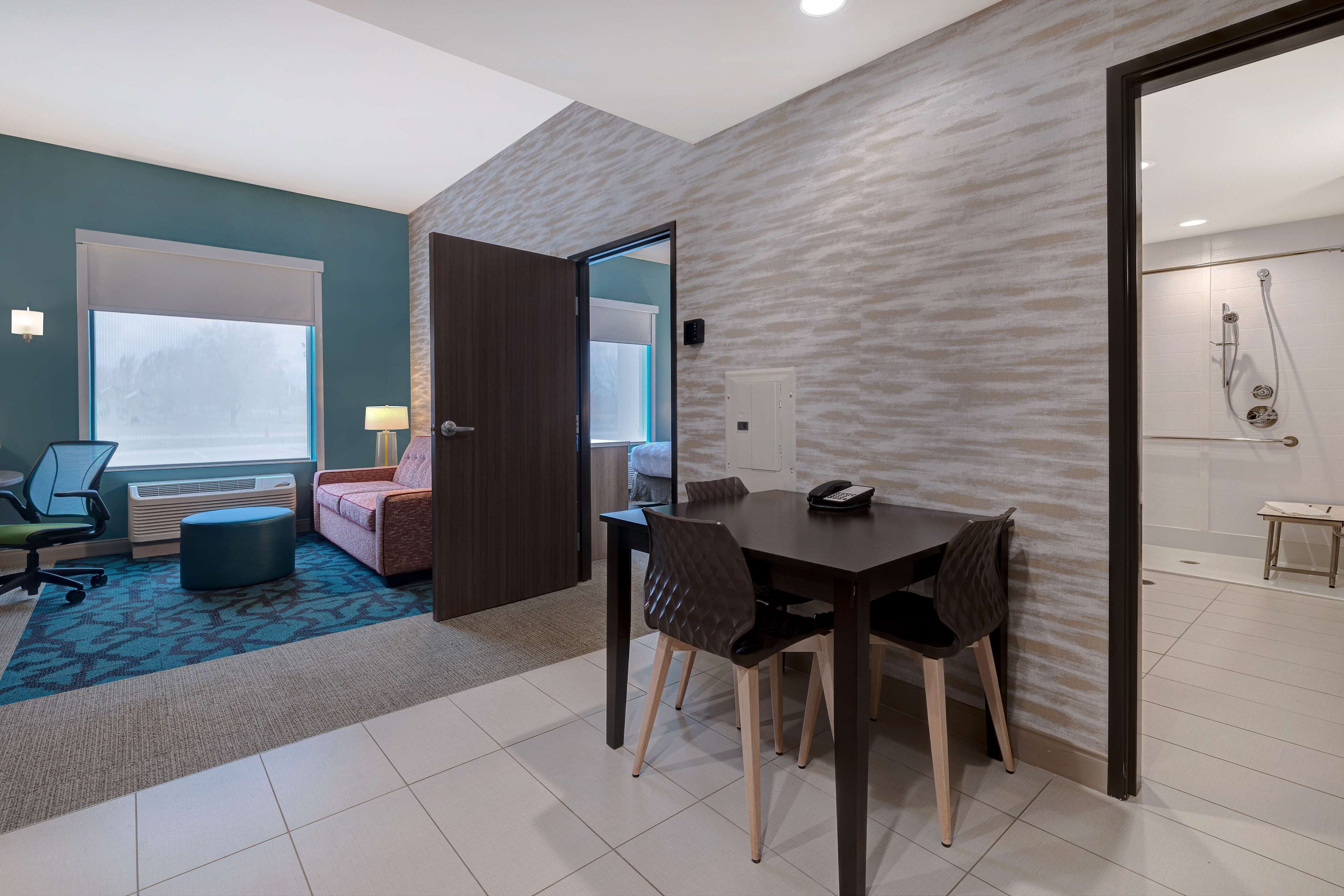 A hotel room with a dining area, a sofa, a chair, a table, and a bathroom visible through an open door.