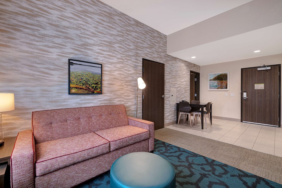 Een moderne hotellobby met een rode bank, een rond blauw voetenbankje, een zithoek met een tafel en stoelen, en een kamerbreed tapijt met dessin.