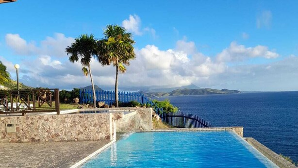 Piscina azul cristalina con una persona nadando, rodeada por un muro de piedra y una valla azul, con vistas a una masa de agua y montañas lejanas.