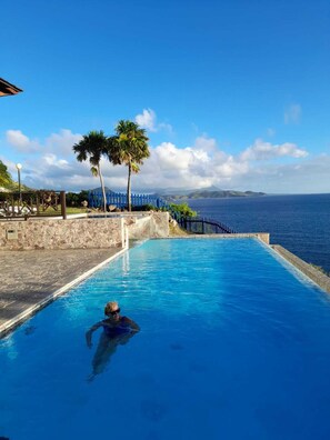 A clear blue pool with a person swimming, surrounded by a stone wall and a blue fence, overlooking a body of water and distant mountains. - Lovely hillside villa overlooking the Caribbean Sea with private plunge pool. (Frigate Bay)