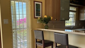 Una cocina con encimera de madera, dos taburetes de bar, un jarrón con flores y una ventana con persianas.