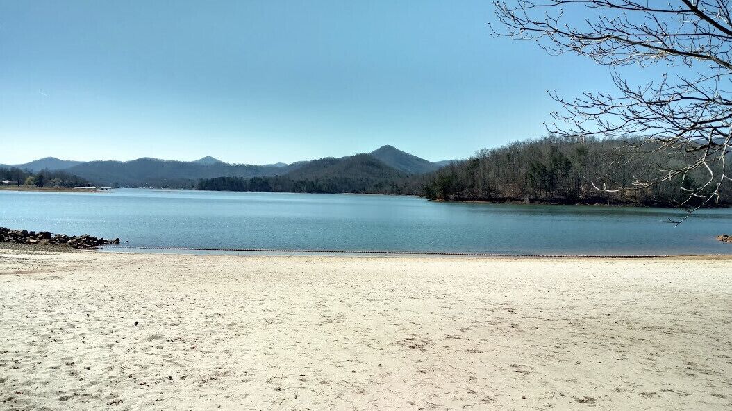 Une plage bordée d’un rivage sablonneux, un lac paisible et, au loin, une chaîne de montagnes boisée.