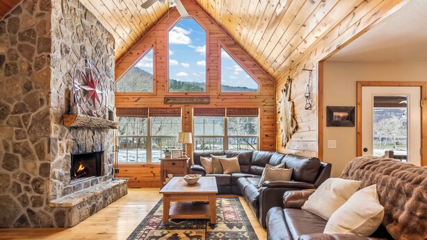 A cozy living room with a stone fireplace, leather sofas, and large windows offering a mountain view. - Riverfront cabin, arcade, hot tub, pool, trout fishing, pet friendly (Hiawassee)