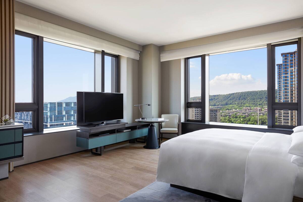 A modern hotel room with a large bed, a flat-screen TV, a desk, and a chair. The room has large windows offering a city view.