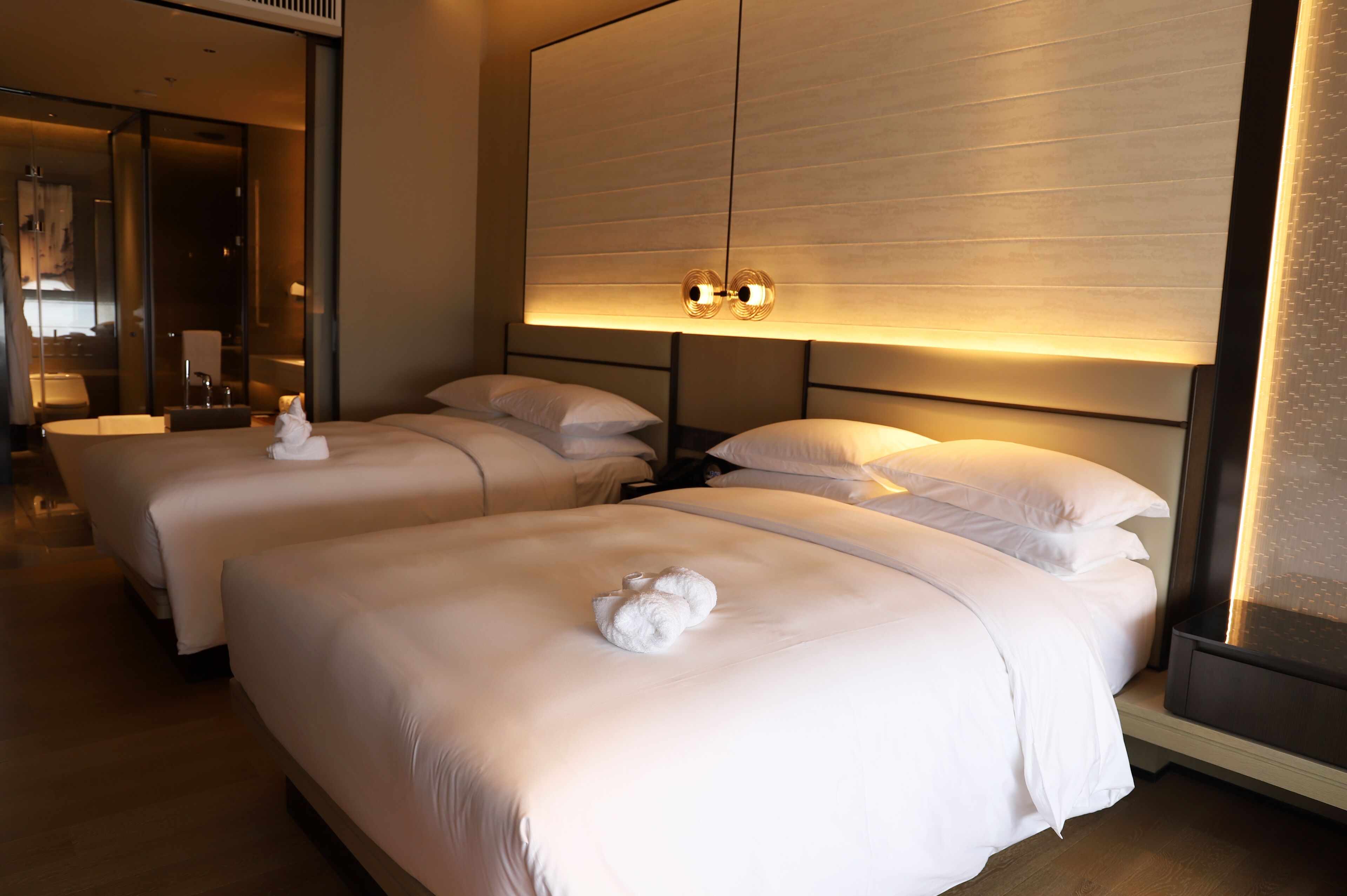 Habitación de hotel con dos camas, cada una con ropa de cama blanca y una toalla sobre la cama. Hay una lámpara grande fijada a la pared sobre las camas.