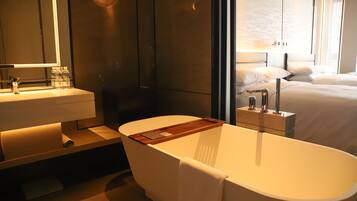 A modern bathroom with a freestanding bathtub, a vanity with a mirror, and a bedside table with a lamp.