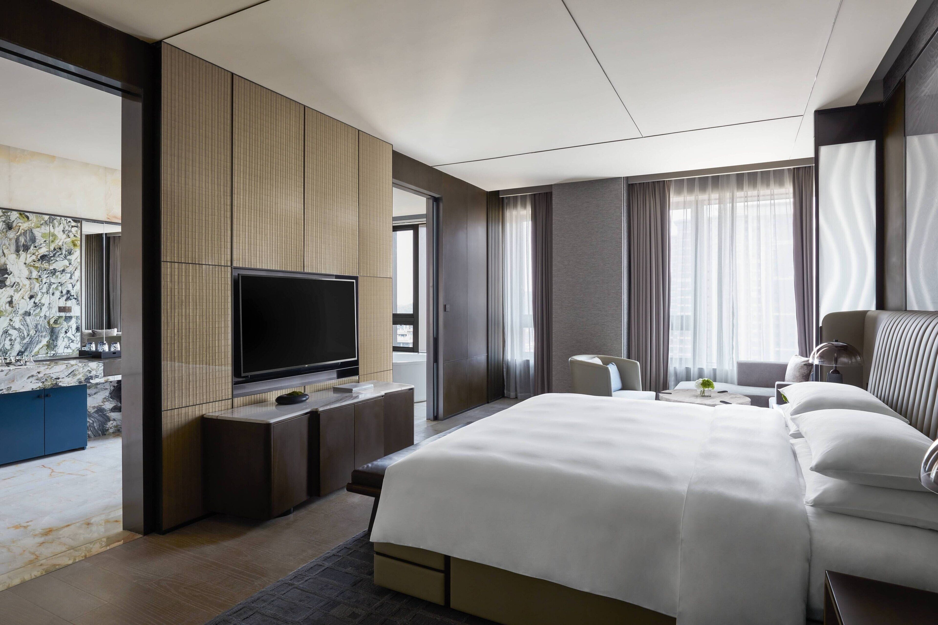 A modern hotel room with a large bed, a flat-screen TV, a desk with a chair, and a small table with a lamp.