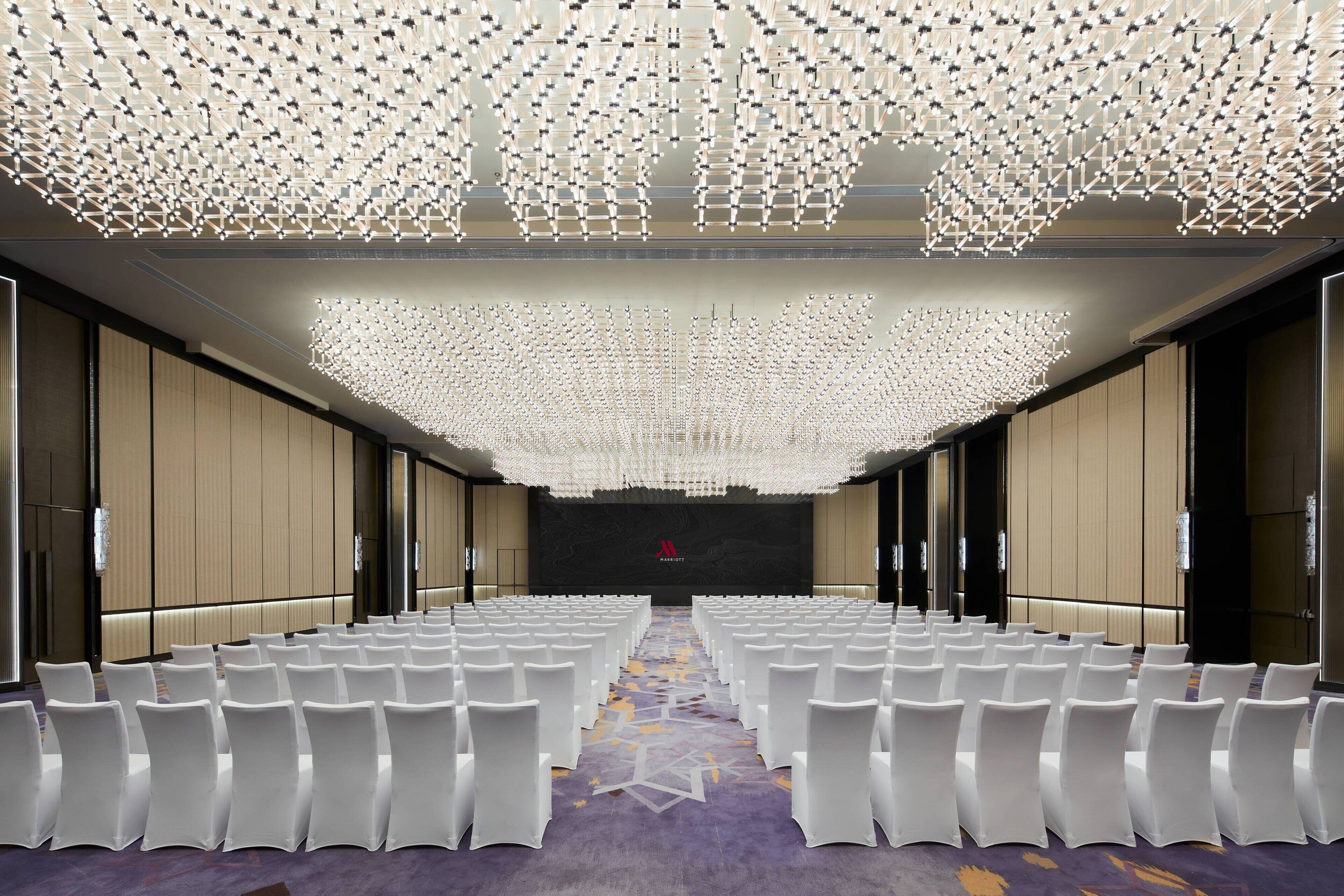 A conference room with rows of white chairs, a patterned carpet, and a ceiling with a geometric light fixture.