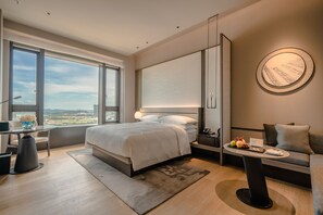 A modern hotel room with a large bed, a desk, a sofa, a round wall decoration, and a view through large windows.