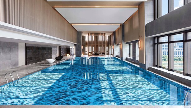 A modern indoor swimming pool with a patterned blue tile floor, large windows, and a view of the cityscape. - Wuhan Marriott Hotel Optics Valley (Wuhan)