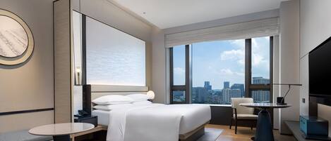 A modern hotel room with a bed, a desk, a chair, and a large window offering a city view.
