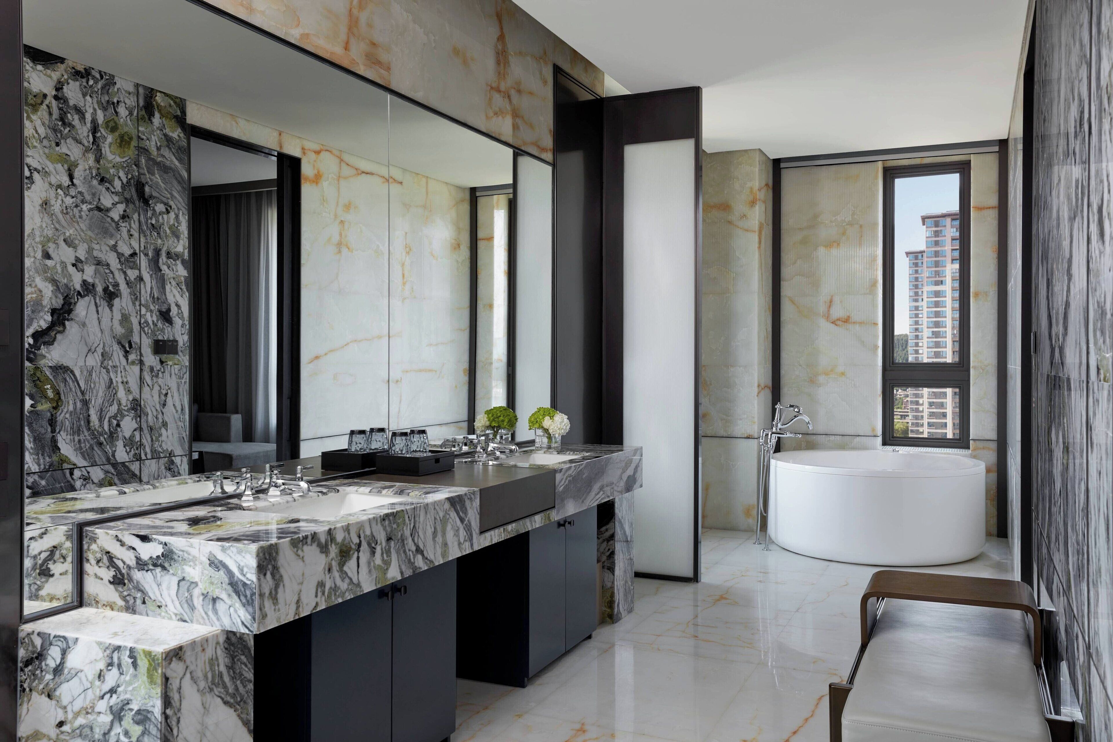 Un bagno moderno con un mobile lavabo in marmo, uno specchio grande, una vasca da bagno indipendente e vista su un edificio attraverso una finestra.