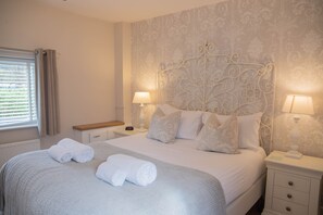 A neatly made bed with pillows and two rolled towels, a bedside table with a lamp, and a window with curtains. - Dale Cottage (Telford)