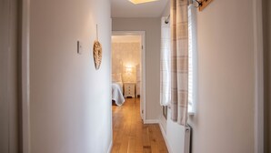 Un couloir étroit avec un plancher en bois, un lit à l’arrière-plan, un radiateur fixé au mur et un luminaire suspendu au plafond.