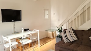 Uma sala de estar moderna com uma TV de tela plana, uma área de jantar com uma mesa e cadeiras brancas, uma pequena mesa com tampo de vidro, um sofá marrom com almofadas listradas e uma escada.
