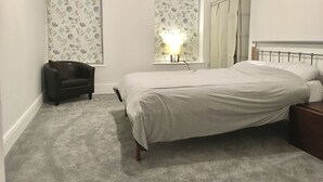 Un dormitorio con cama, una silla, una mesita de noche, un espejo y una ventana con estampado floral.