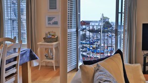 A room with a view of a harbor, a dining table with chairs, a small white table with a basket of books, and a sofa with a patterned cushion. - Harbourside Apartment (St Austell)