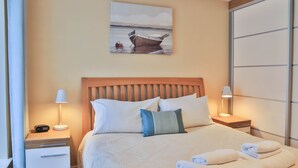 A neatly made bed with white and blue pillows, a wooden headboard, and two bedside lamps. - Harbourside Apartment (St Austell)