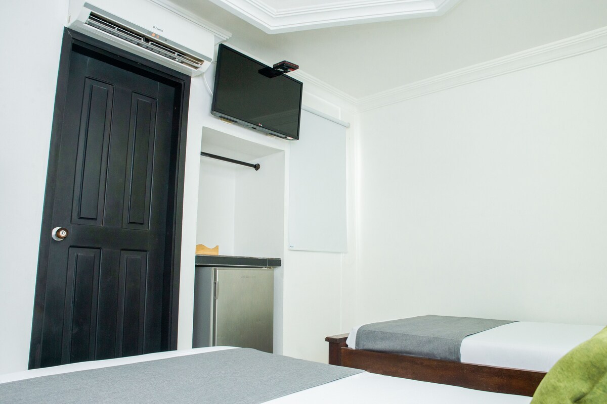 A hotel room with a dark wooden door, a flat-screen TV mounted above it, a small refrigerator, and a bed with a grey blanket.