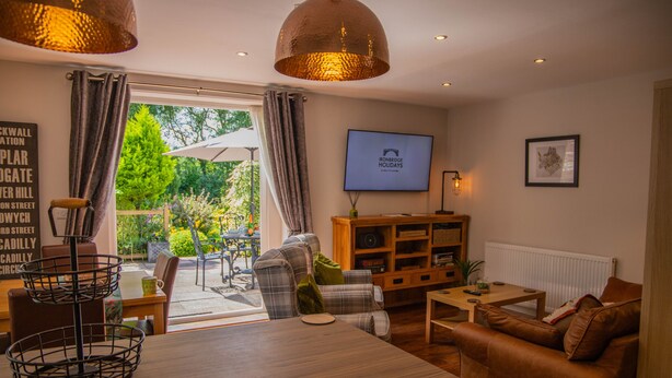 A cozy living room with a sofa, a coffee table, and a TV mounted on the wall. There is a bookshelf and a window with curtains. - River Mist (Telford)