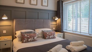 A neatly made bed with decorative pillows, a bedside table with a lamp, and a window with blinds. - River Mist (Telford)