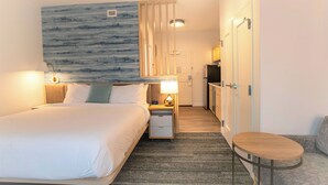 A hotel room with a bed, bedside table with lamp, a small round table, a kitchenette, and a bathroom with a shower. - TownePlace Suites by Marriott Conroe (Conroe)