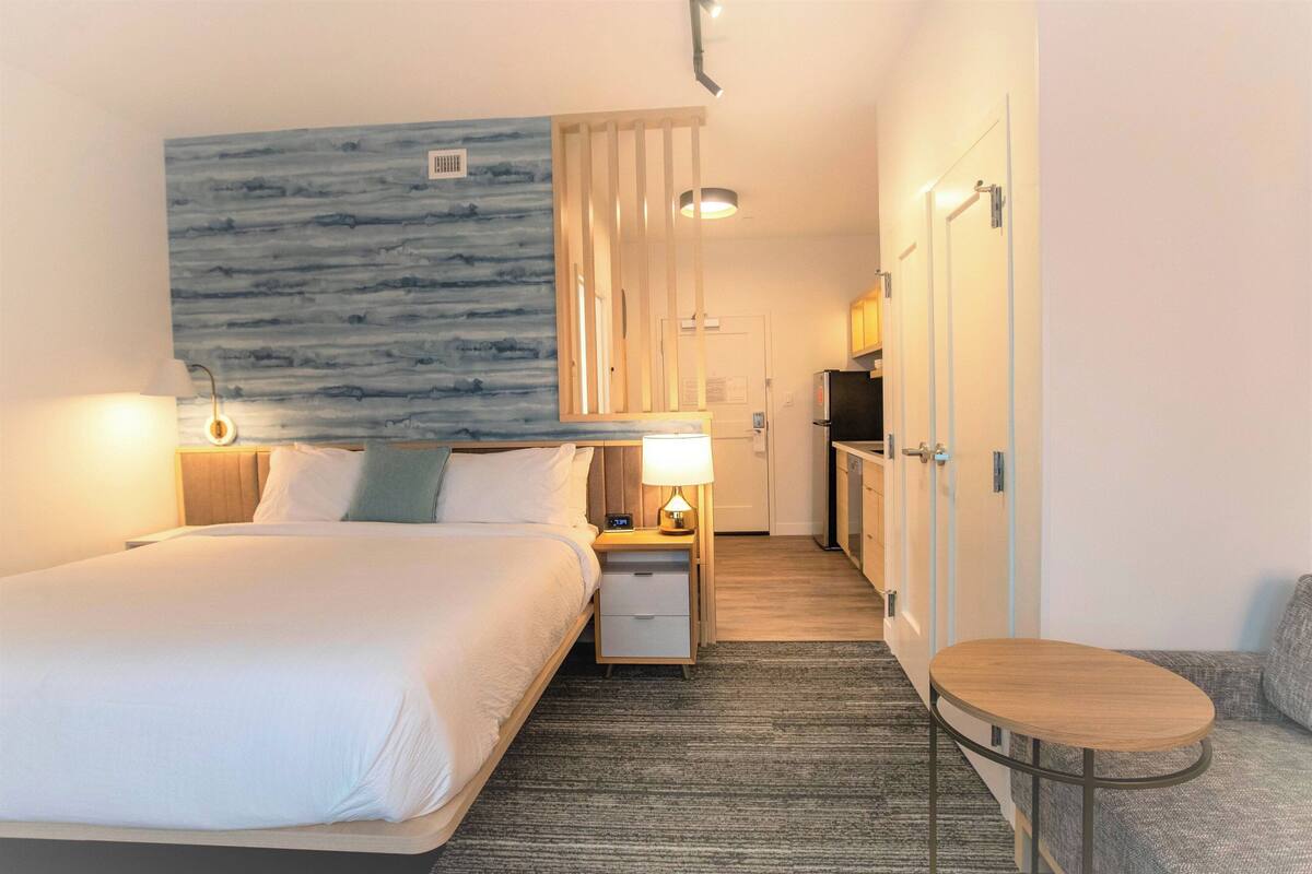 A hotel room with a bed, bedside table with lamp, a small round table, a kitchenette, and a bathroom with a shower.