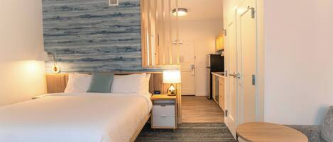 A hotel room with a bed, bedside table with lamp, a small round table, a kitchenette, and a bathroom with a shower.
