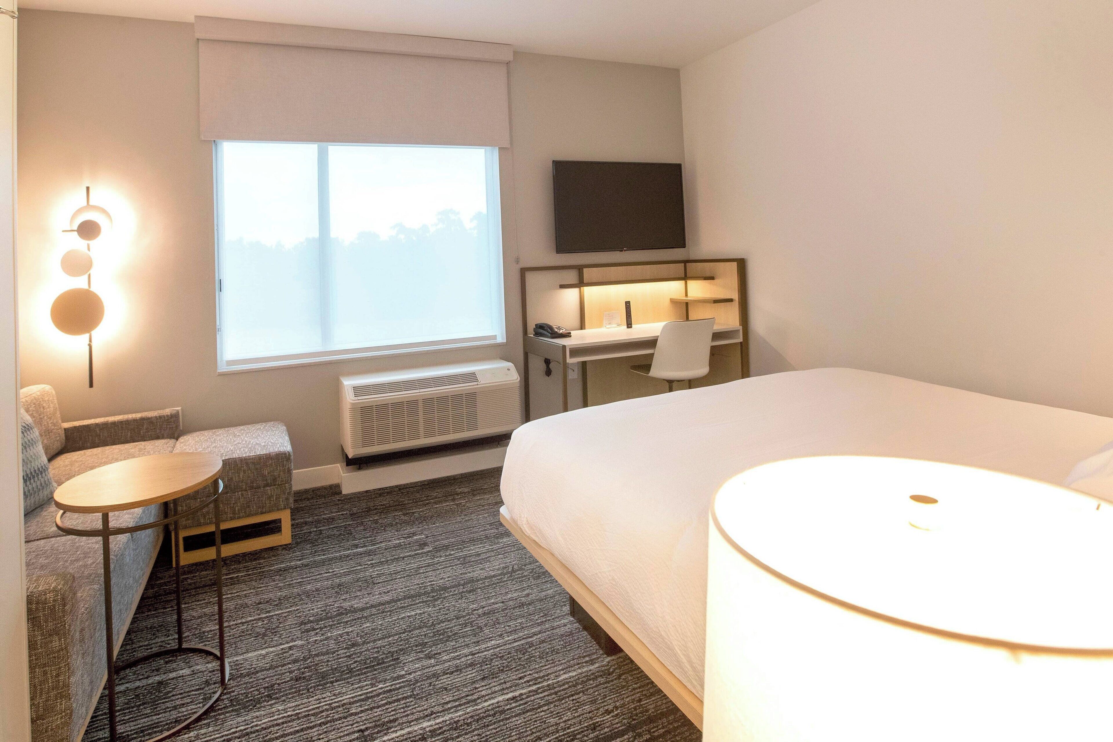 A hotel room with a bed, a desk with a chair, a small table, a TV, and a window with a view.