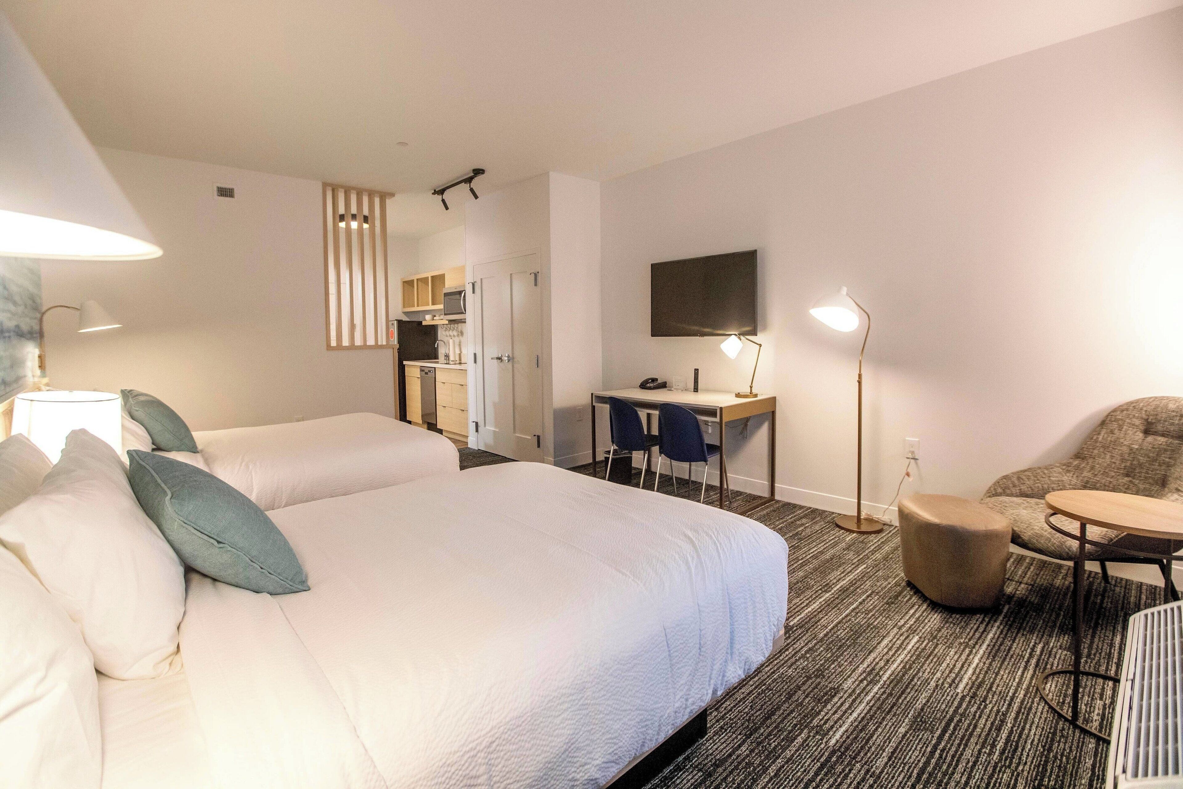A hotel room with two beds, a desk, a chair, a TV, and a lamp.