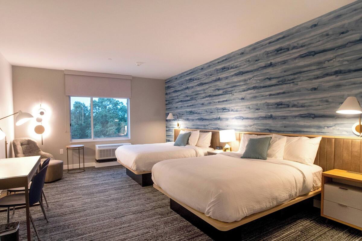A hotel room with two beds, a desk, and a chair. There is a large window with a view of trees.