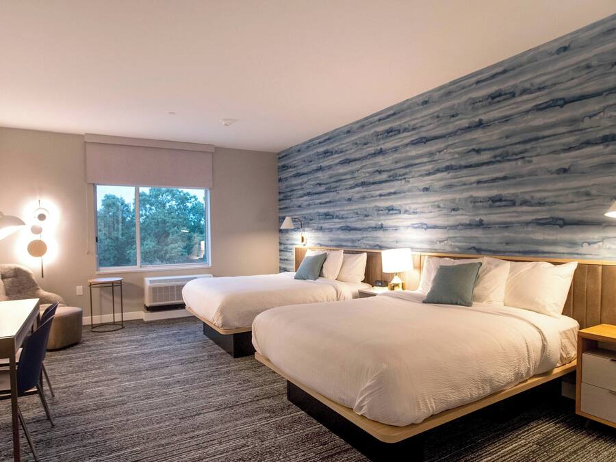 Habitación de hotel con dos camas, un escritorio y una silla. Amplia ventana con vistas a árboles.