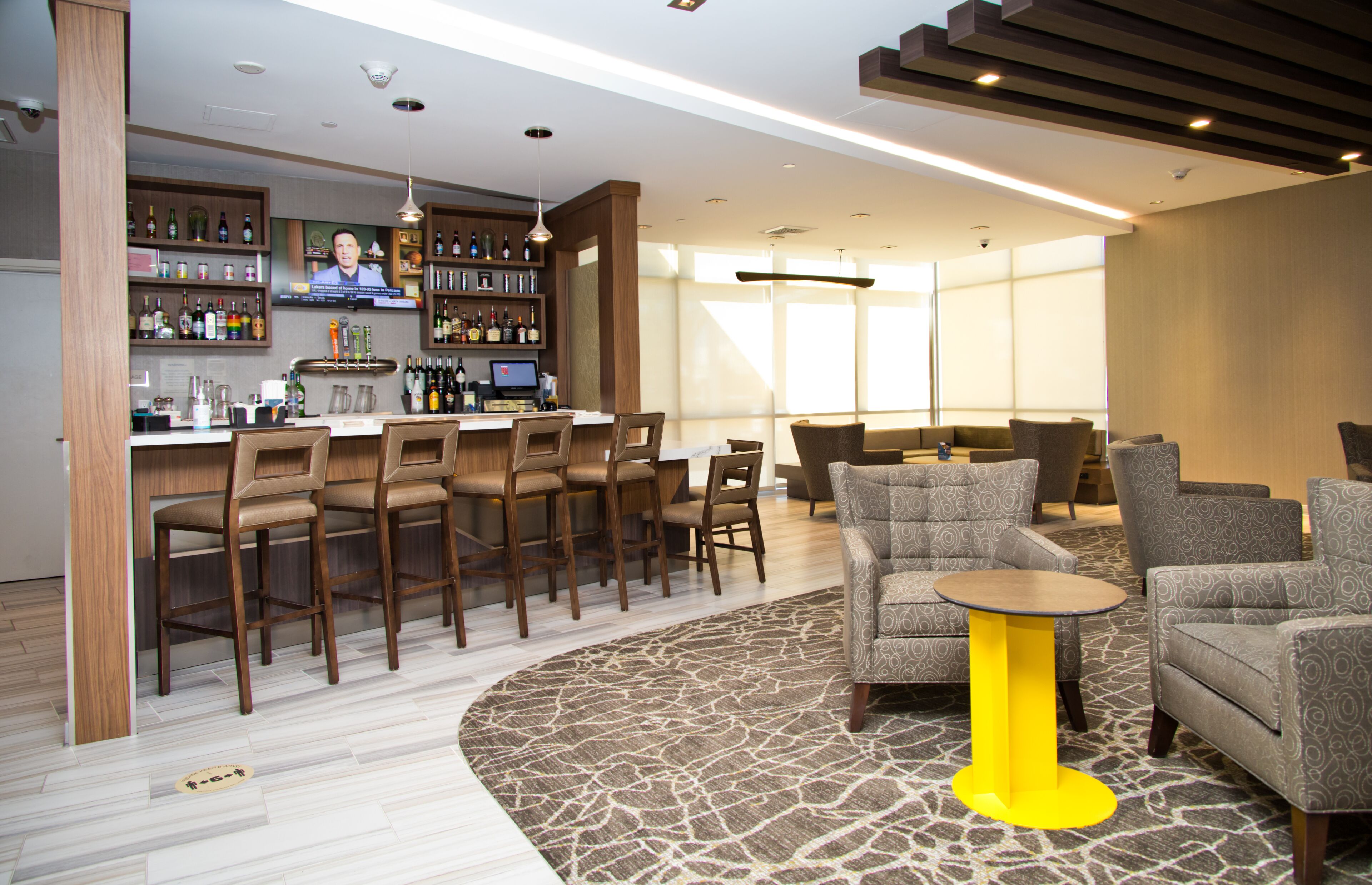 A modern bar area with a television screen, bar stools, and a variety of bottles. There is a seating area with sofas and a patterned rug.