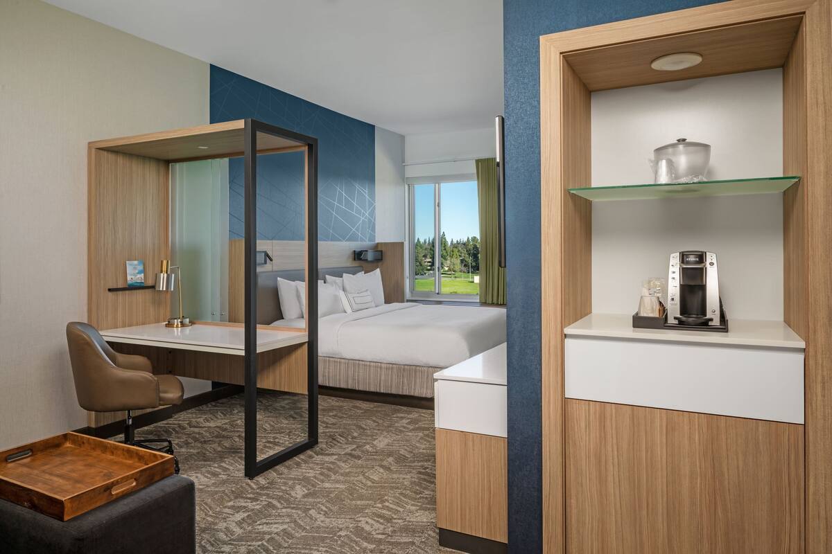 A hotel room with a bed, a desk, a chair, a coffee maker, and a shelf with a teapot.