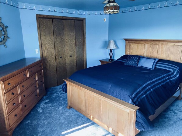 A bedroom with a wooden bed frame, a dresser, a nightstand with a lamp, and a door with a nautical-themed wall decal.