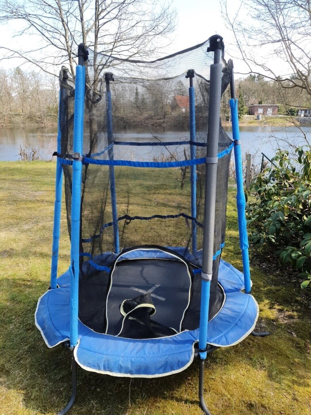 A trampoline with a blue safety net enclosure, set on grass near a body of water.