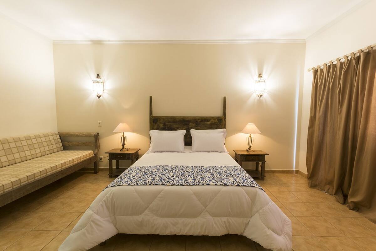 Ein Schlafzimmer mit einem Bett, zwei Nachttischlampen, einem Holzkopfteil, einem beigefarbenen Sofa und einem Vorhang.